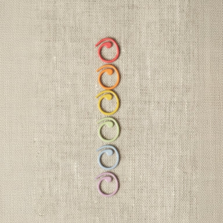 Seks farverige plastikloops formet som spiralors, arrangeret lodret på beige stof.